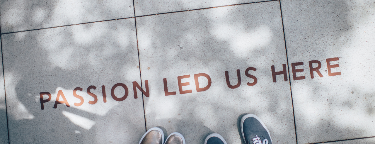 two sets of feet in sneakers stand on a sidewalk, near lettering that reads "Passion led us here"
