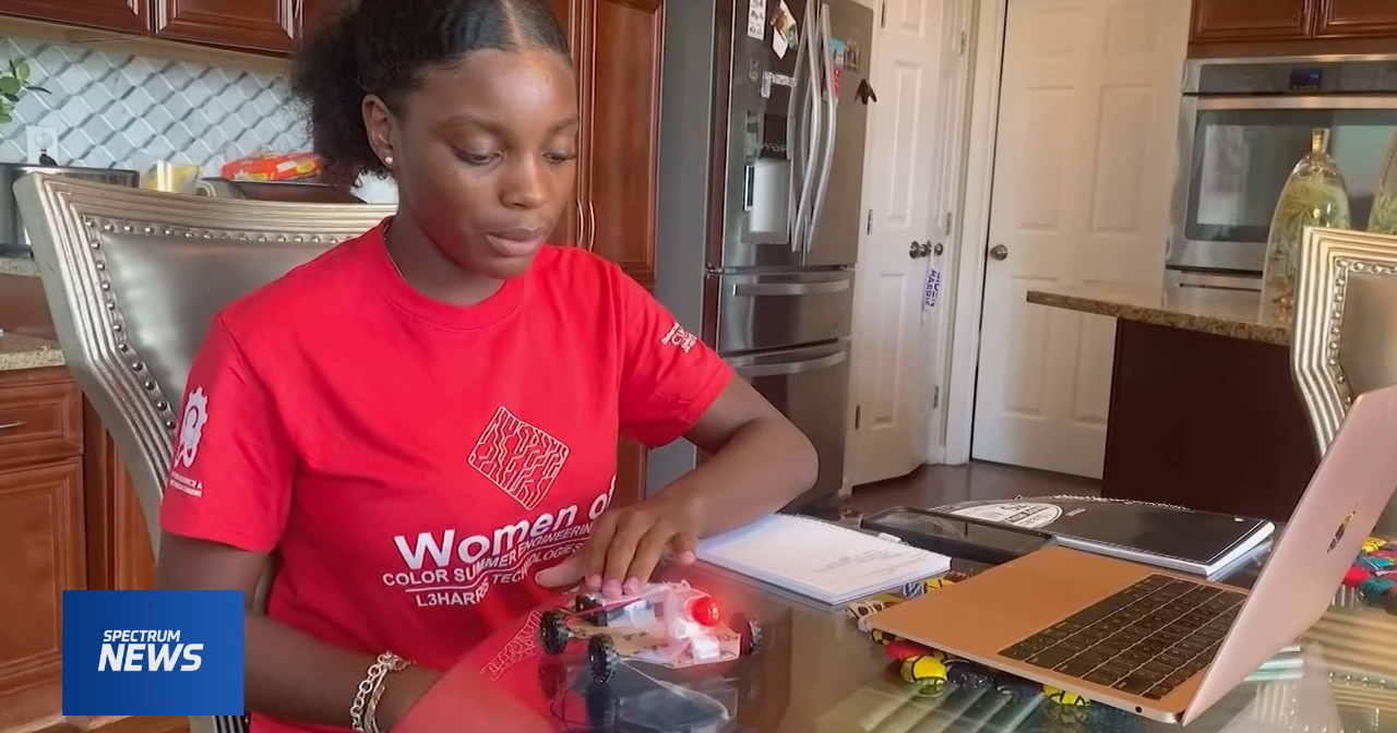 Mariah Couch sits at a table in front of a tiny wheeled robot and an open laptop.