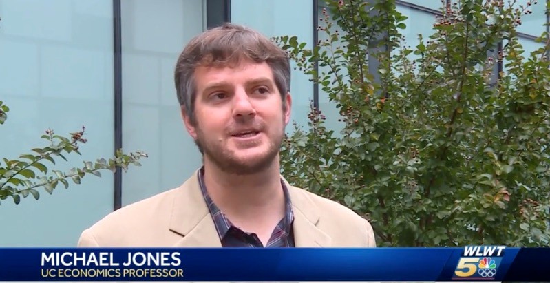 A man in a jacket answers a question from a television reporter.