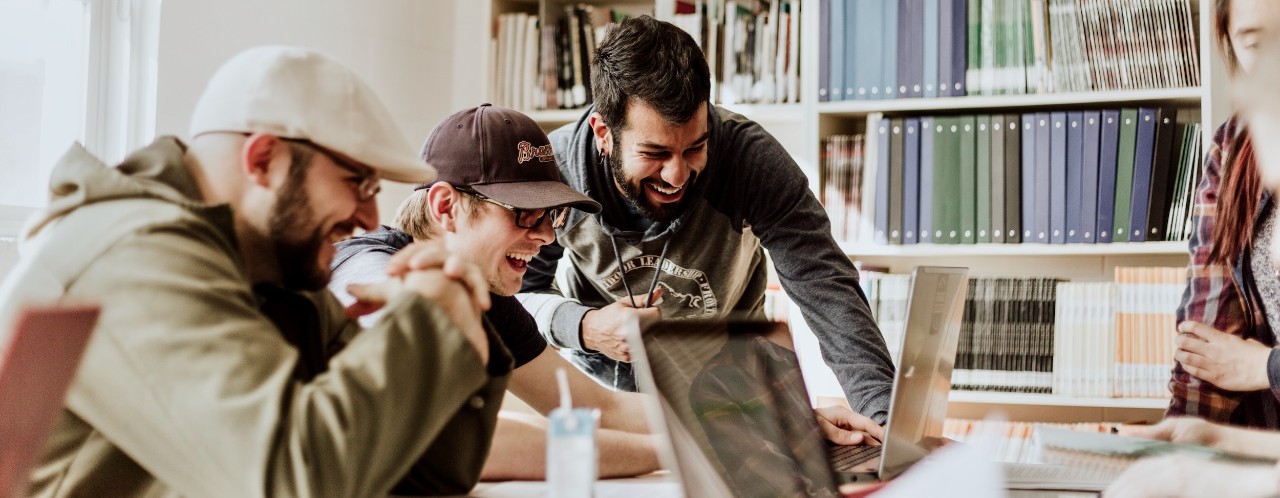 Three male students sit at table near bookshelves laughing at screens on two laptops