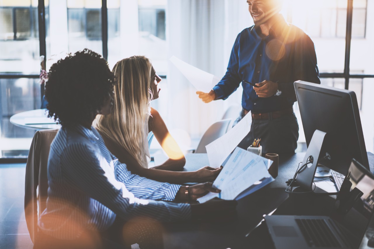 Young team of coworkers making great meeting discussion in modern coworking office.Hispanic businessman talking with two beautiful womans.Teamwork process.Horizontal,blurred background,sun effect