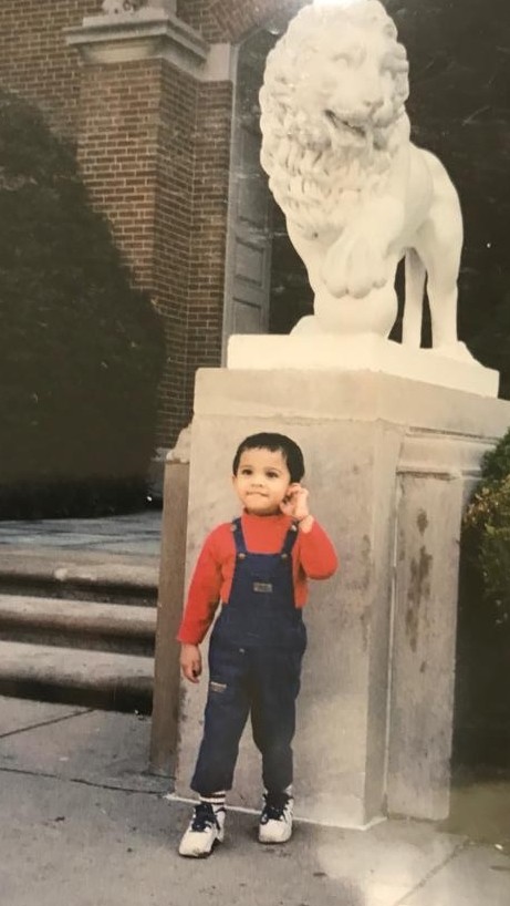 Chinmay, a toddler, stands in front of the lion statue in front of the College of Arts & Sciences