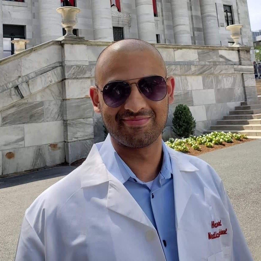Chinmay Bakshi stands in front of a building at Harvard Medical School wearing his white coat