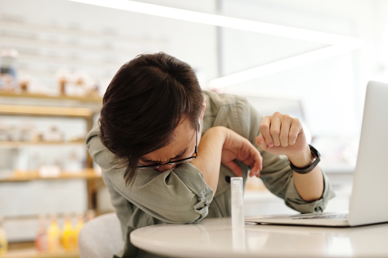 a photo of a man sneezing into his elbow sitting at a desk