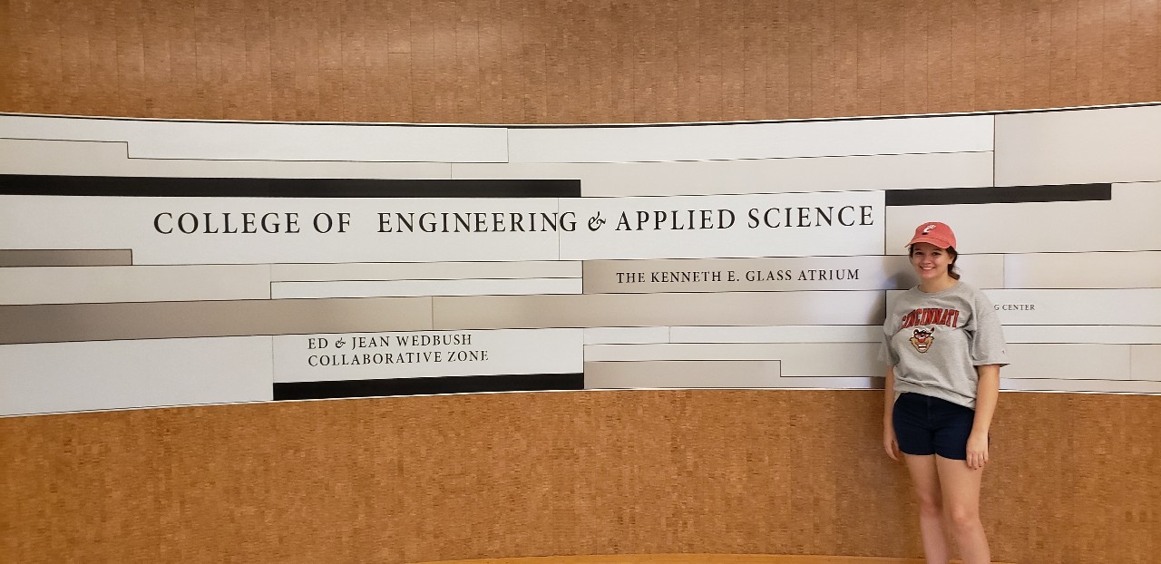 Taylor Daugherty posing in front of the College of Engineering and Applied Sciences sign and wearing Bearcat t-shirt and hat
