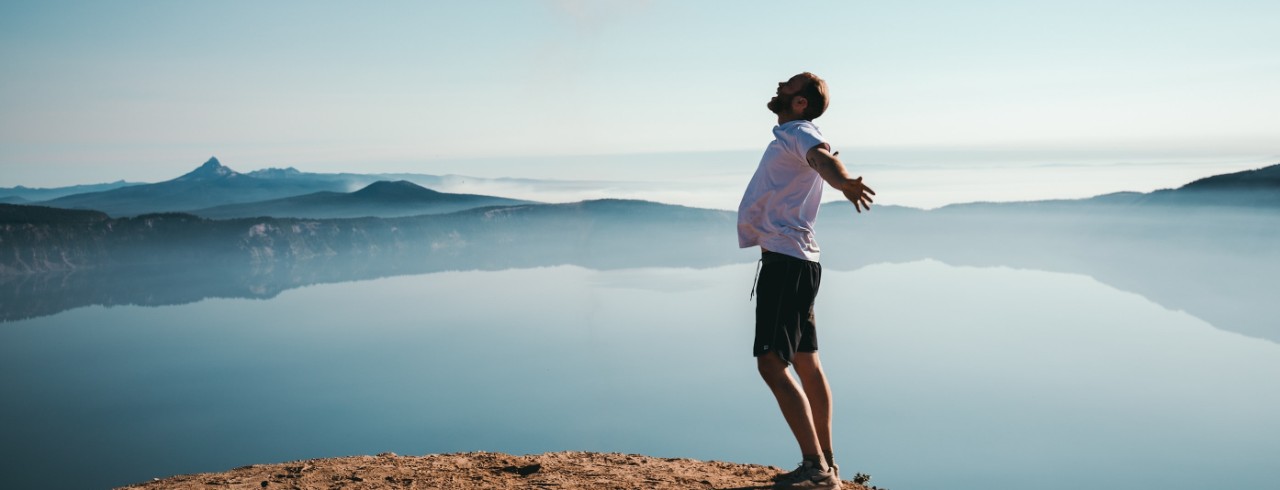 Man stands with his arms outstretched and head up on the shore of a body of water