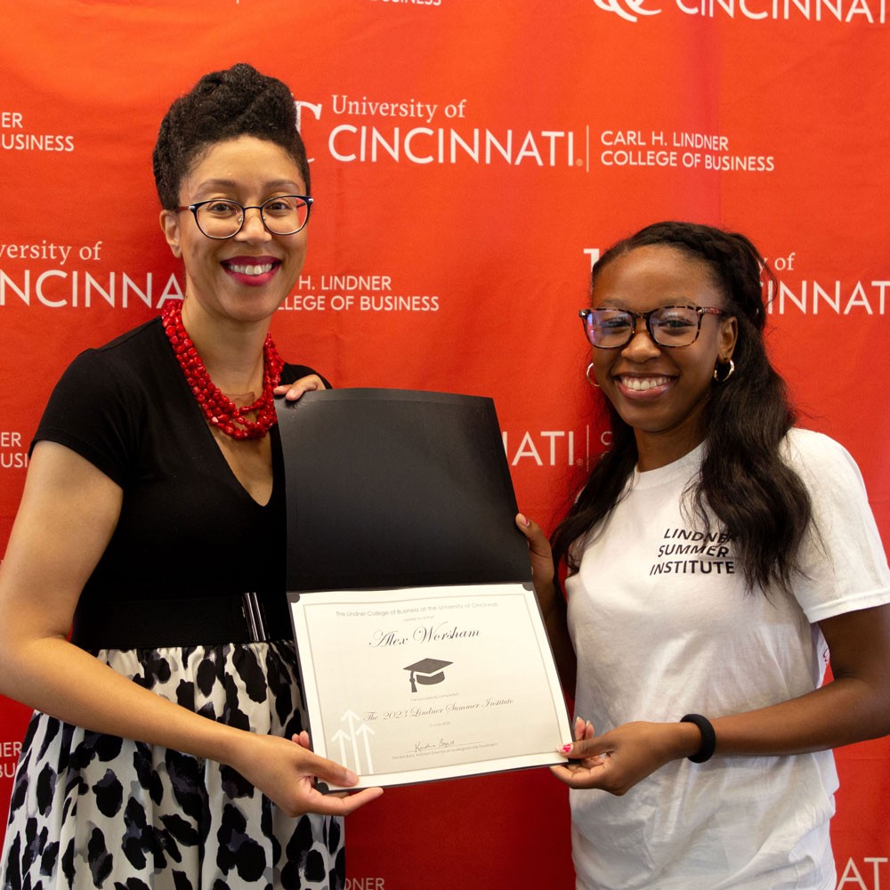 woman wearing black top and red necklace and glasses presents white certificate in black folder to a female high school student wearing a white shirt and glasses against a red backdrop