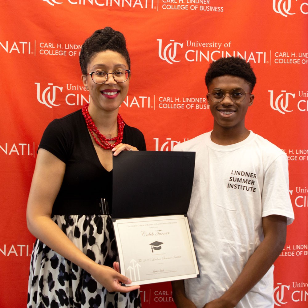 woman wearing black top and red necklace and glasses presents white certificate in black folder to a male high school student wearing a white shirt and glasses against a red backdrop