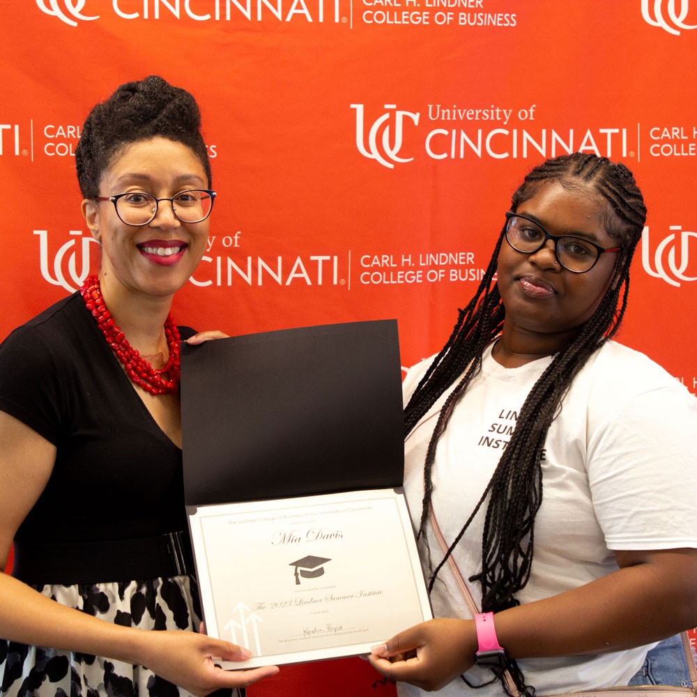 woman wearing black top and red necklace and glasses presents white certificate in black folder to a female high school student wearing a white shirt and glasses against a red backdrop