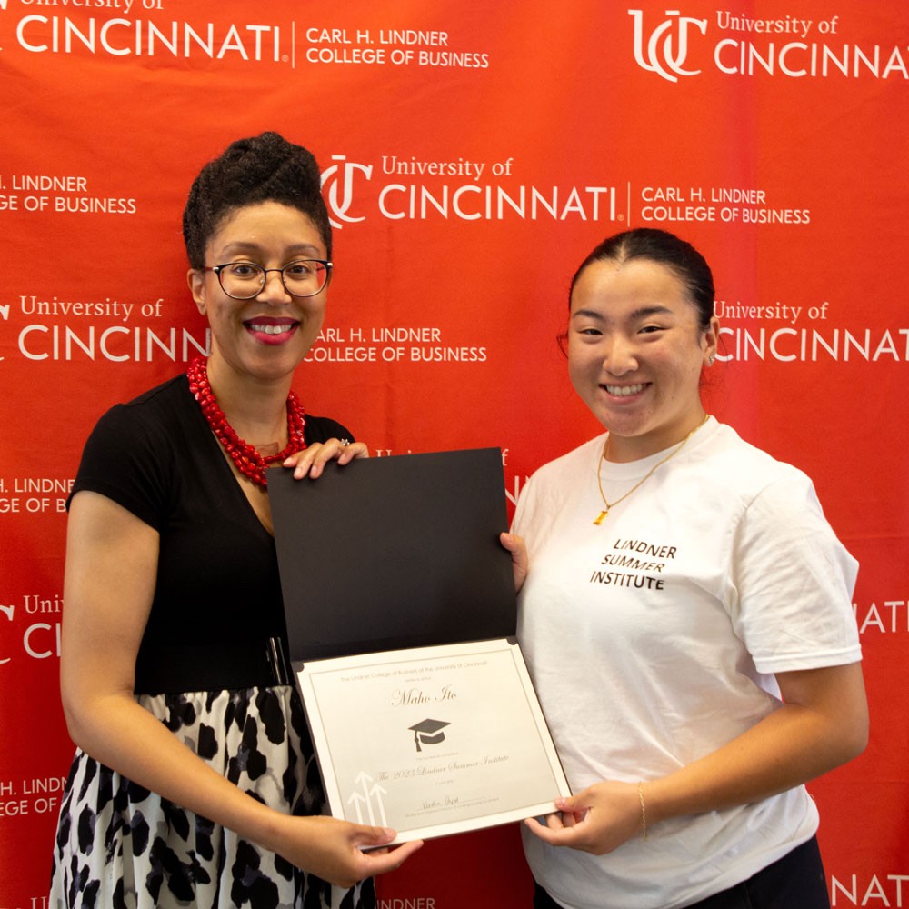 woman wearing black top and red necklace and glasses presents white certificate in black folder to a female high school student wearing a white shirt against a red backdrop