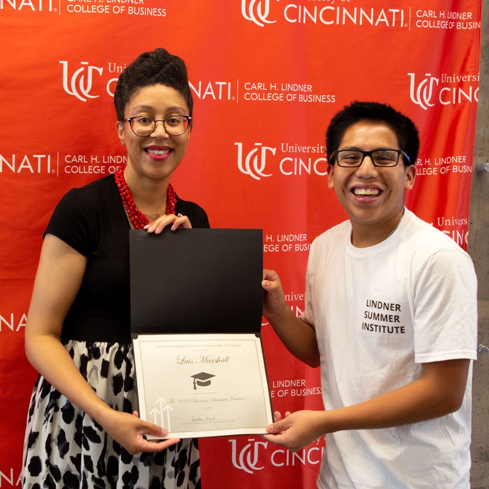 woman wearing black top and red necklace and glasses presents white certificate in black folder to a male high school student wearing a white shirt and glasses against a red backdrop