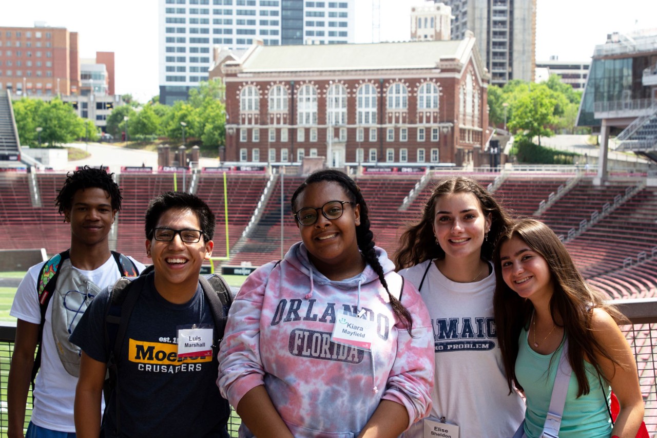 five students wearing multicolor t shirts stand in front of a fence overlooking the football field and bowl at Nippert Stadium