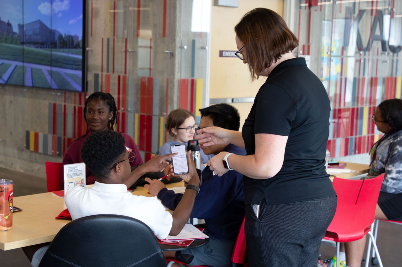 a group of five students sit at a rectangular table writing in notebooks or on their phones and the student in the foreground is holding their phone up to a woman wearing a black polo with her back to us to view something on the phone