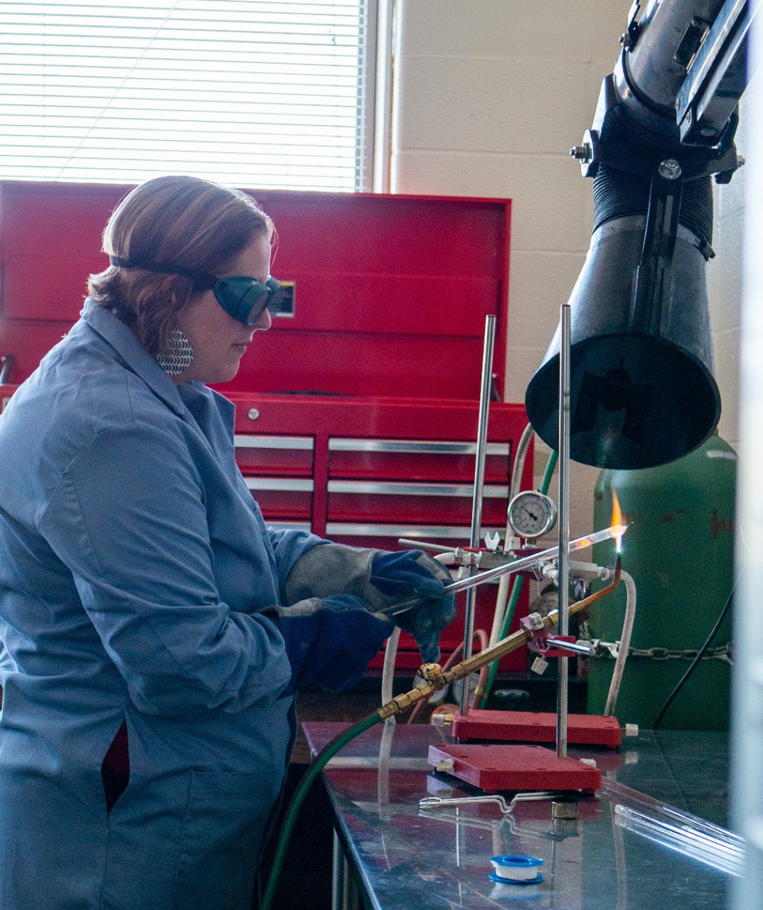 Woman in a blue lab coat, protective goggles and gloves holds metal over a flame in a lab