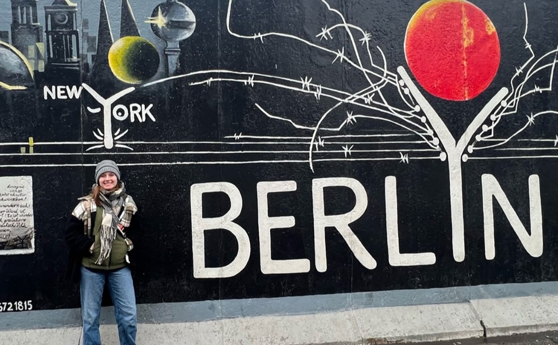 A young woman poses in front of a street mural, predominantly black and reading in large letters “Berlin” and in small letters “New York” and “Tokyo.” Part of the mural shows a stylized cityscape, and some equally stylized planets.