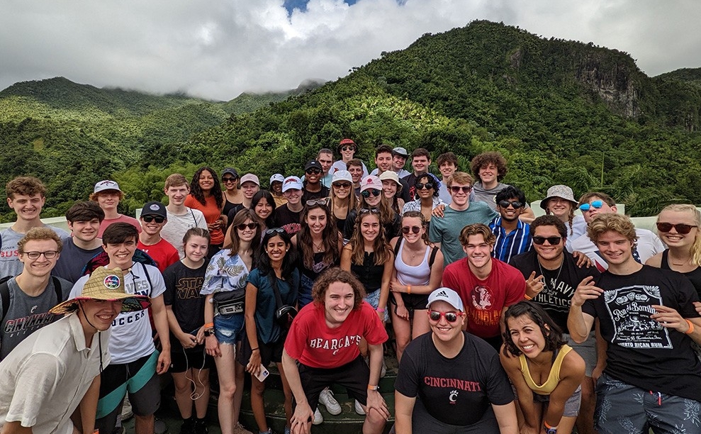 A group, some wearing UC spirit wear, pose on a hilltop with deeply forested mountains in the background, topped by clouds.