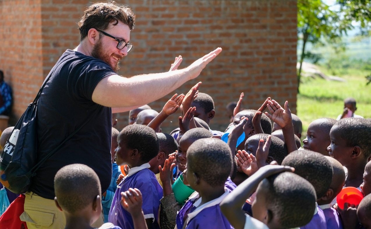 Elliott, wearing a UC t-shirt, raises his hands to high-five Tanzanian children in a group surrounding him. They are much smaller than he is. Some raise their hands to return the gesture; others stare.