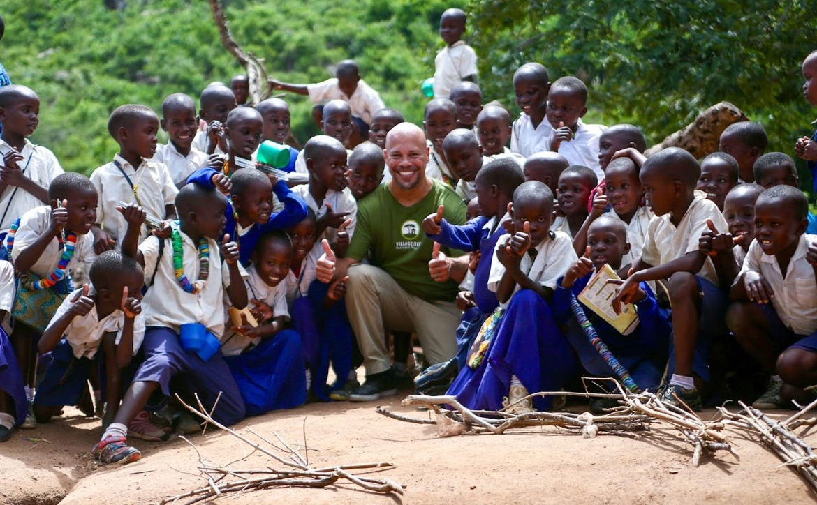 Dr. Lewis poses with a large group of children in blue and white uniforms on a rock in a forested landscape.