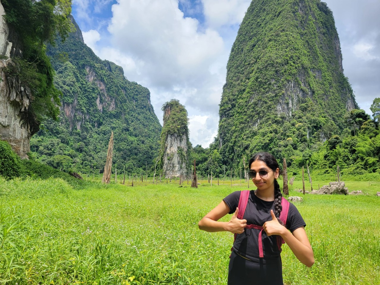Prisha Vatsal, a young woman wearing a backpack, gestures thumbs up. She is standing on a field in front of massive stone formations covered with lush greenery.