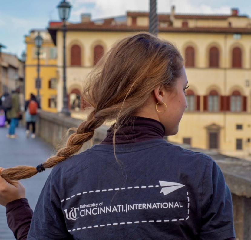 A young woman stands on a bridge with her back to the camera. She is holding her long braid away from her back so that the logo on her t-shirt, which reads “UC University of Cincinnati|International” is visible to the camera. Beyond her, the bridge, its rail topped with lamp posts, stretches toward some yellow stucco buildings.