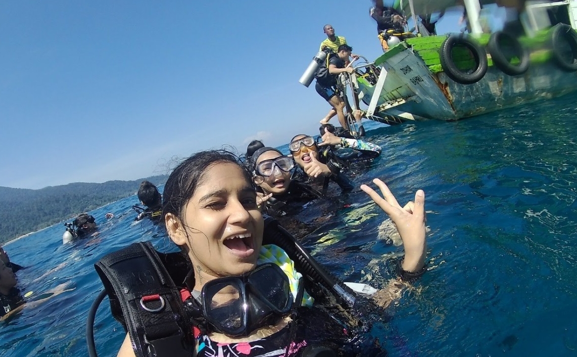 Three women wearing dive gear and treading water gesture for the camera; behind them are other divers and the boat they are diving from, and in the background, mountains are visible.