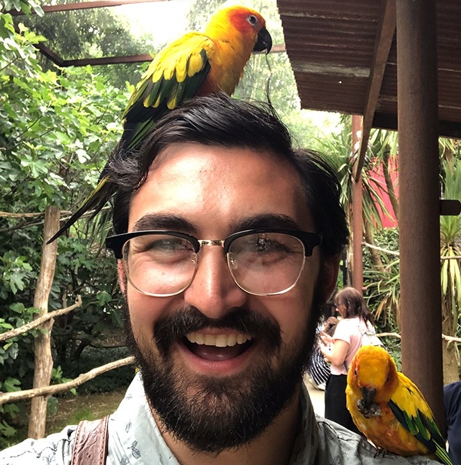 A broadly smiling man stands on a covered wooden porch while brightly colored birds perch on his head and shoulder.