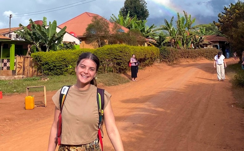 A young woman in a backpack stands on a dirt road. A few people are on the road behind her. Small buildings behind hedges and palm trees are in the background, and a rainbow is visible in the sky.