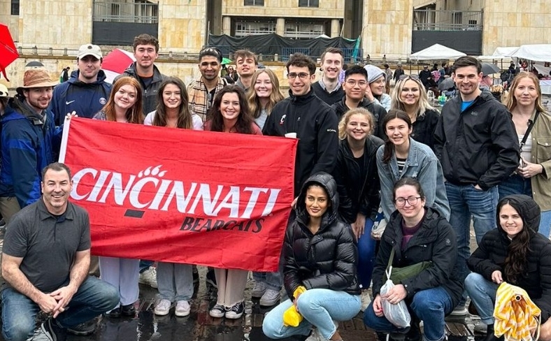 A group of people pose in the rain in a plaza before a yellow stone building. They hold a banner reading “Cincinnati Bearcats.”