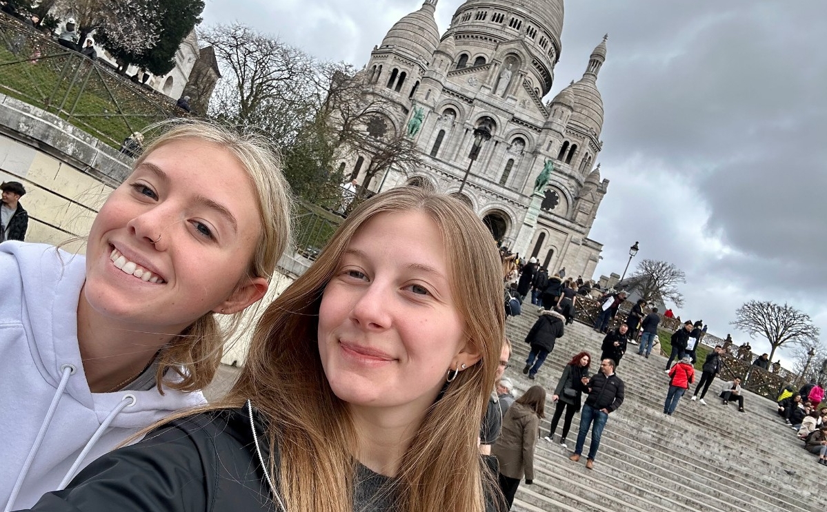 Two young women take a selfie in a plaza before a long, wide flight of steps; in the background behind them, the Sacre Coeur basilica in Paris is visible.
