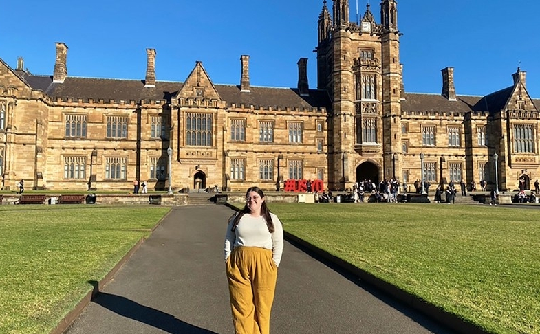 A young woman stands on the walkway in the quadrangle of the University of Sydney, with a golden stone building in traditional architecture behind her. 