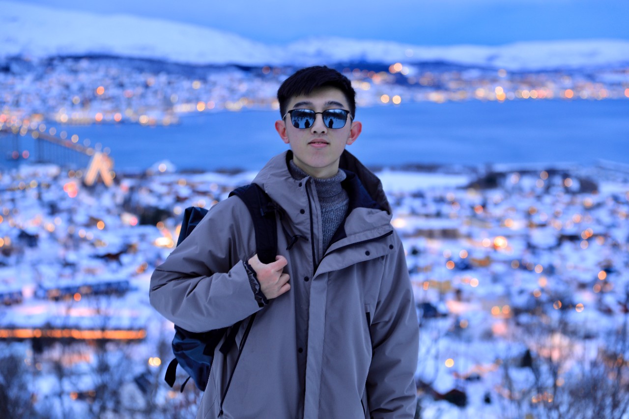 A young woman in a yellow hardhat and a UC International t-shirt smiles up at the camera from an archaeological dig site in Pylos. She is clearing an area of the ground with a small brush.