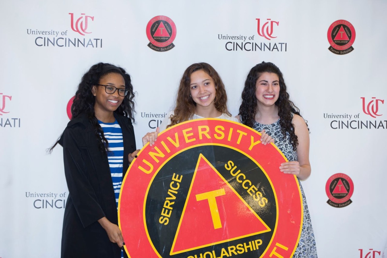 Three women hold a large sign with the logo of UC's Darwin T. Turner Scholars Program.