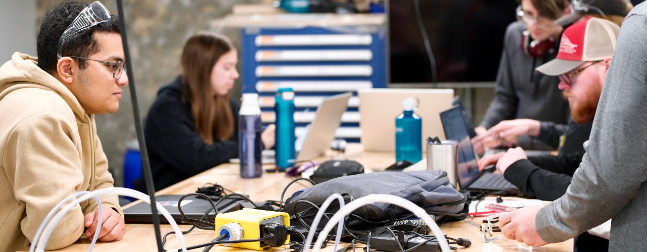 Several students sitting at makerspace workshop table surrounded by wires, tools and laptop.