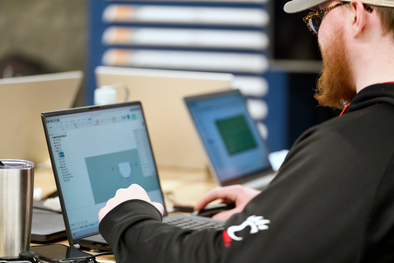 Male college student wearing baseball cap sits at a table working on a laptop.