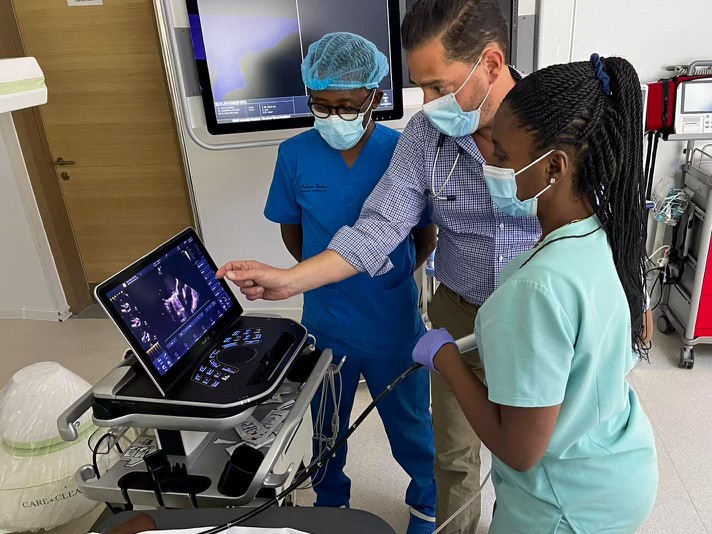 Rubinstein points out an image on a laptop to two other people who are masked and gloved and wearing scrubs; the setting appears to be in a hospital, and other electronic equipment is visible