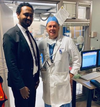 Alt-text: Meshesha and Hattemer pose together; Hattemer is wearing surgical scrubs and a white UC lab jacket. There is medical equipment in the room beside them.