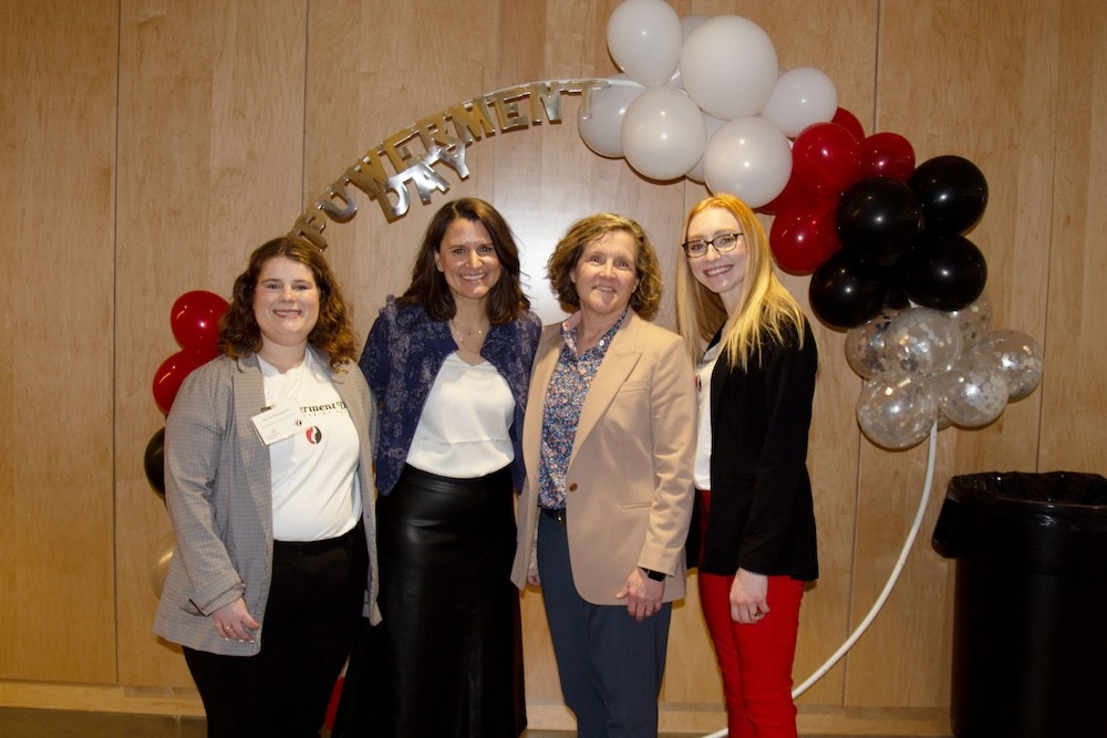 Four women in business professional and business casual attire stand in front of a balloon backdrop that says "Empowerment Day" and wood panel wall