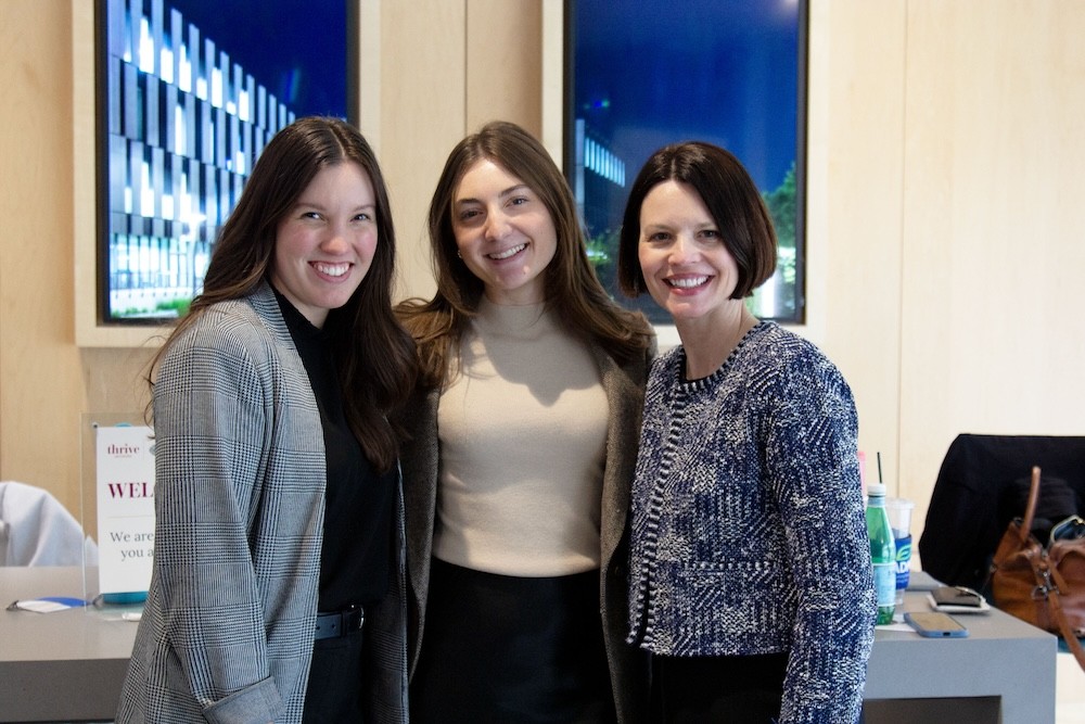 three women with dark hair and business professional attire stand as a group and smile in front of their company table in the Lindner Hall atrium