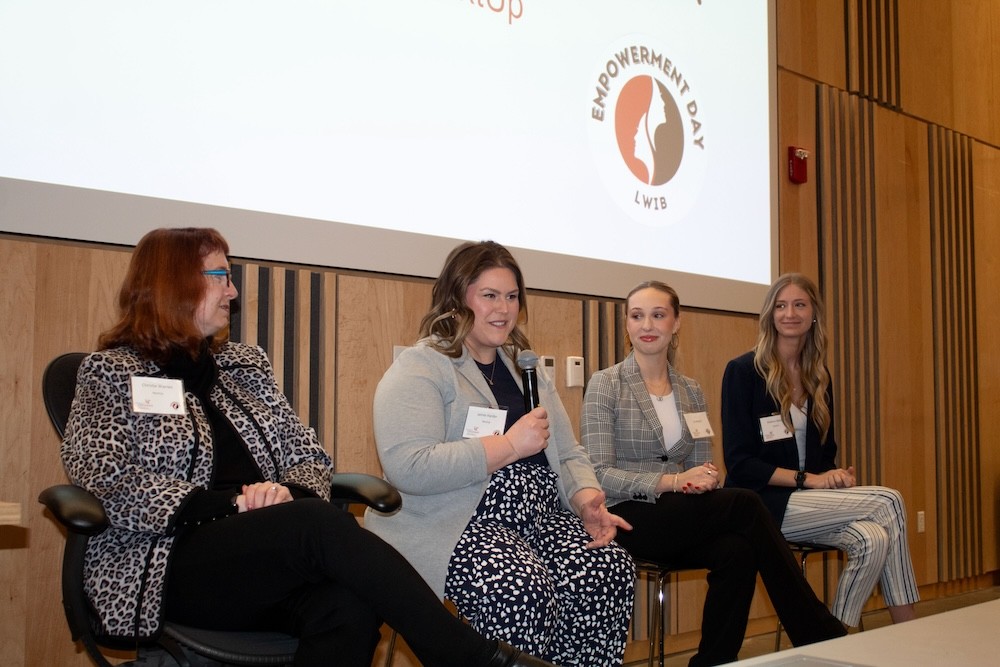 Four women in business casual attire sit on high chairs in front of a classroom. The second one from the left is holding and speaking into a microphone