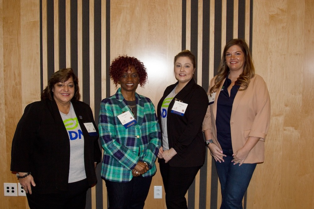 Four women in business attire stand in front of a wood panel wall