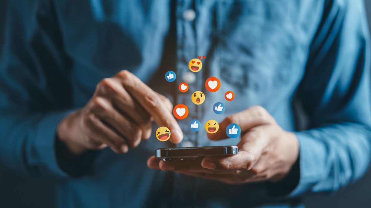 A man scrolls a social media app on his phone with reaction emojis coming out of the screen.