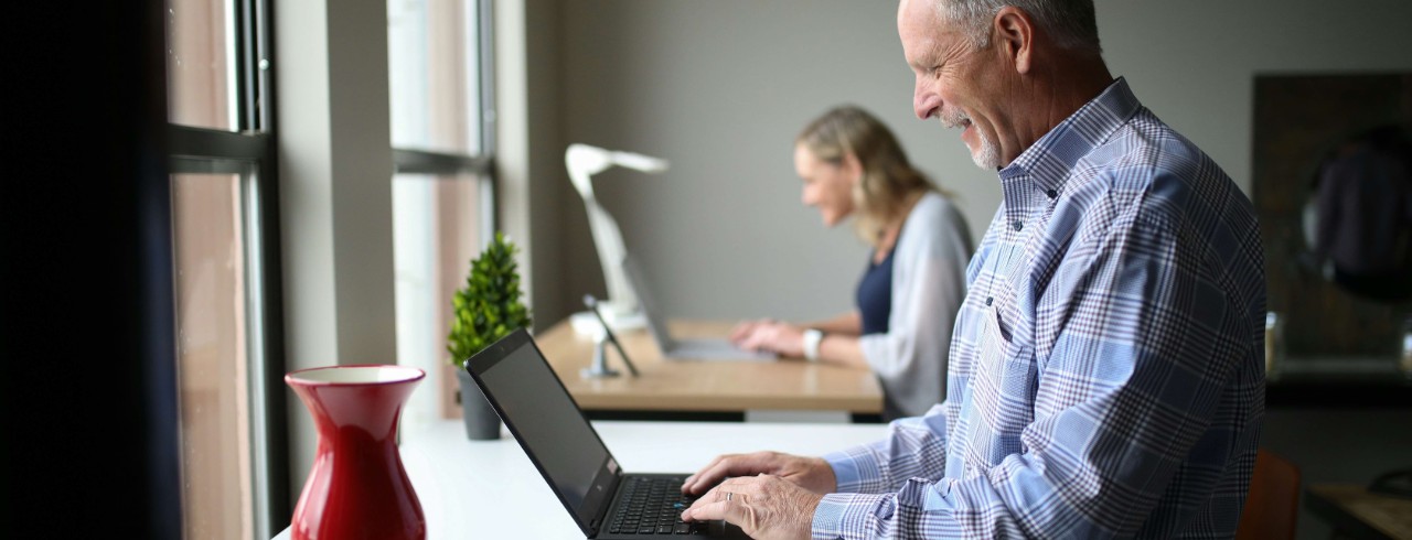 A man works on a laptop with a woman working on a laptop in the background.