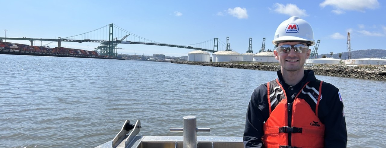 person wearing a hard hat and goggles in a boat in a harbor with oil refining facilities in background