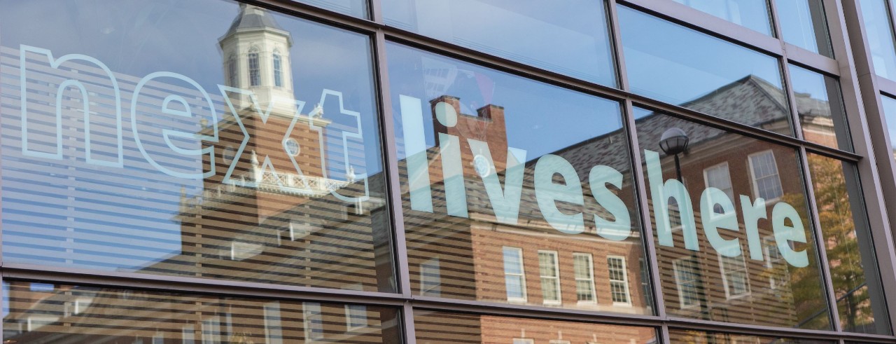 Reflection of UC's Arts and Sciences Hall in a window that has a decal that says "next lives here"