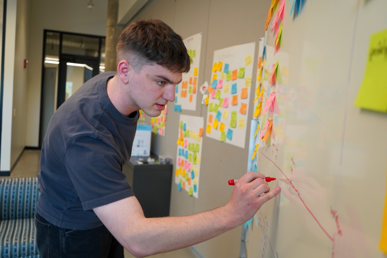 Max Kemats writes on a whiteboard with a red dry erase marker. There are many colorful sticky notes covering the whiteboard.