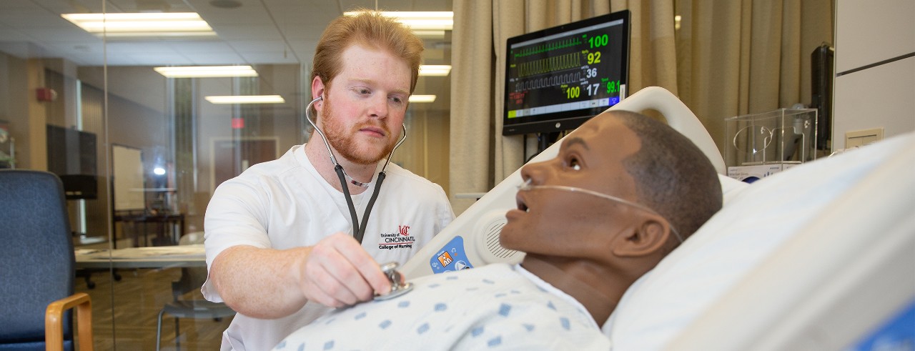 Jack Patterson shown checking the vitals of a mannequin laying in the bed in the UC College of Nursing simulation center