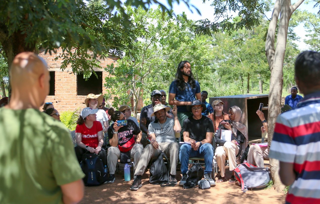 UC student Jonathan Raj stands among other UC students at an outdoor Tanzanian elementary surrounded by trees and leaves.