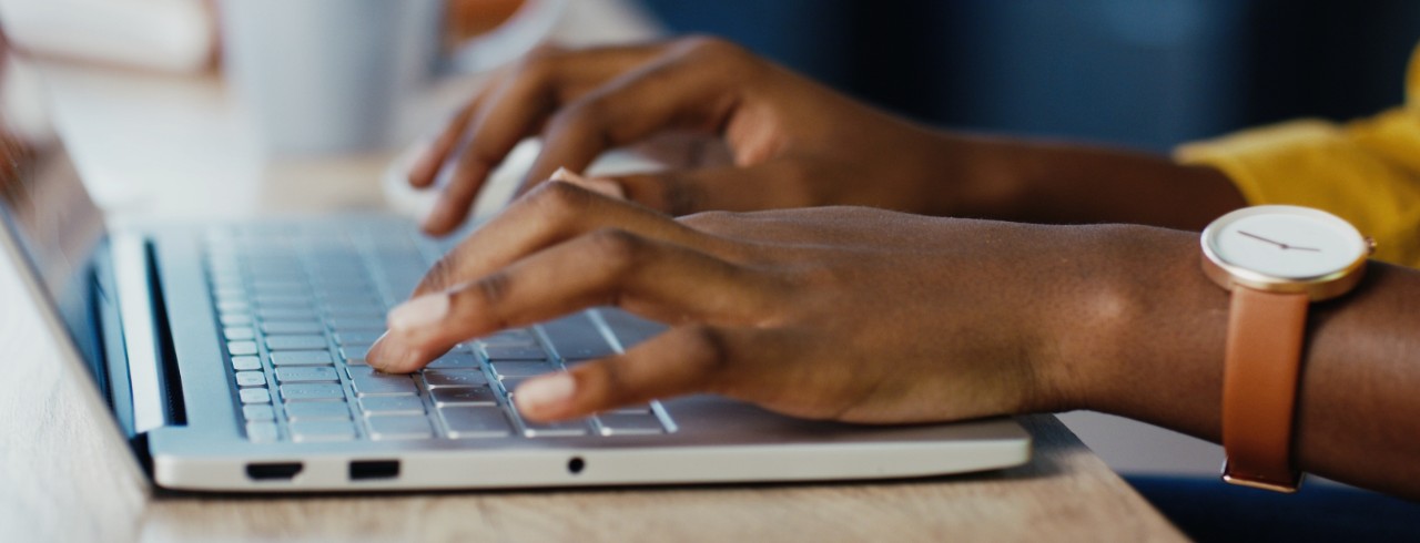 Close up shot of African American female hands typing on laptop while sitting at office desk indoors. Woman fingers tapping and texting on computer keyboard while working in cabinet. 
