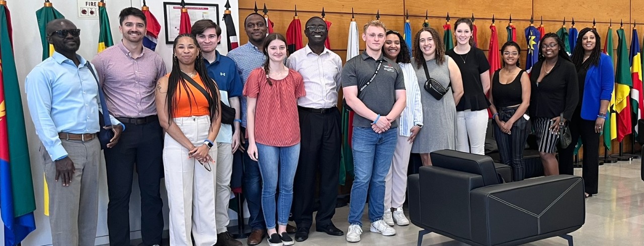Fourteen individuals arranged in a line pose in front of a light wood wall with several nation's flags on poles behind them