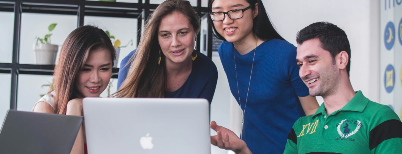 Four people  in an office environment gather around a laptop and smile.